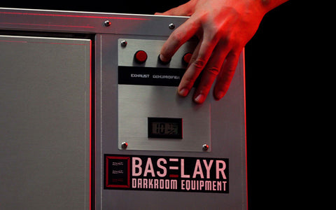 Una mano presiona un botón en un gabinete de secado junto a una calcomanía que dice "Baselayr Equipment Darkroom"
