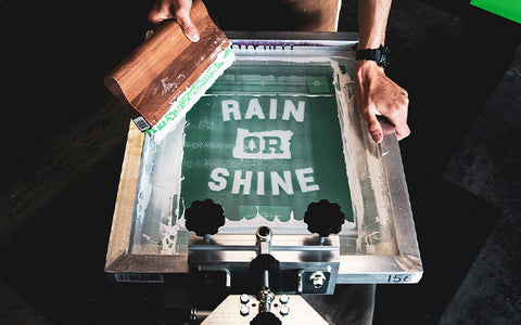 a printer holds a squeegee against a screen reading "rain or shine" in white ink