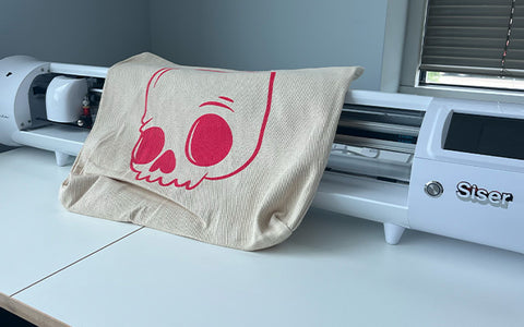 a tote bag printed with a pink skull design rests on a Siser Romeo vinyl cutter and plotter