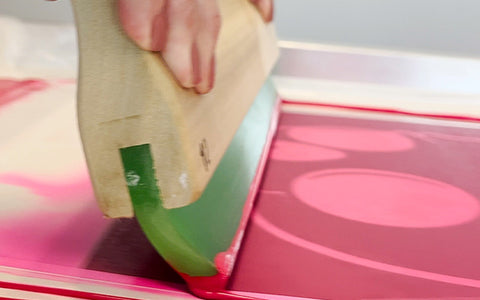 a squeegee pulls pink ink down a screen with a skull design done in vinyl