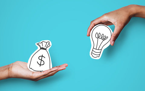 two hands hold paper cutouts, one of a lightbulb and the other a bag of money