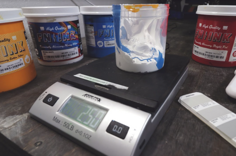 a scale with a container on it with white and blue ink in it. yellow, mixing white, blue, and red ink around it