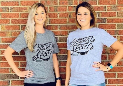 Two women wearing "Support Local" shirts in front of a brick background