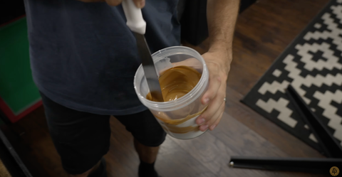 A hand holds an ink spatula and stirs gold/orange ink in a container