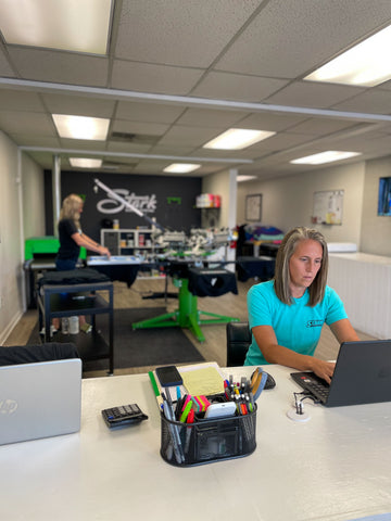 A woman working on a laptop, with another printing on a screen printing press.