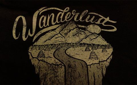 Una camisa negra con una lectura de estampado vintage dorado "Wanderlust" con una escena de montaña y arroyo debajo de ella