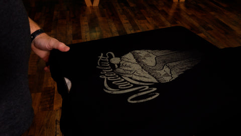 A black shirt with a gold design reading "wanderlust" and showing a mountain scene on a platen. A hand holds the corner of the shirt.