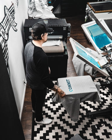 A man wearing a hat stands at a press with a shirt reading "Commit." A black conveyor dryer sits nearby.