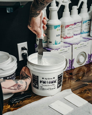 A hand stirs a bucket of white ink with a spatula