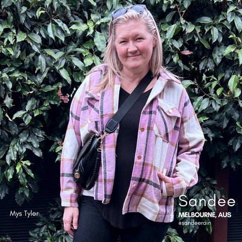 Sandee Rain in a mauve plaid shacket in front of a wall of greenery, wearing a saddle bag crossover body bag