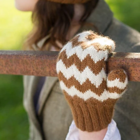 Woman wearing a hand knit gold and white chevron mitten holding on to a wooden fence rail.