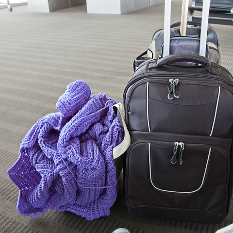 Suitcase and bag with purple crochet blanket project sitting on the floor at an airport.