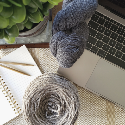 Computer open with a blue skein and naturally colored cake of yarn with notebook and knitting needles.