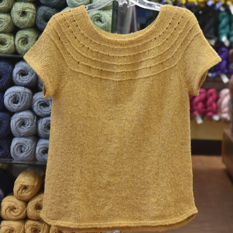 Yellow short-sleeved sweater hanging in front of wall of yarn.