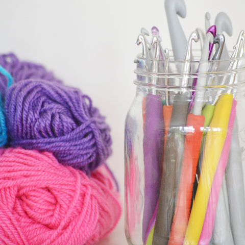 Pink and purple yarn next to a jar of brightly colored crochet hooks.