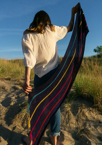 Woman facing away from the camera holding a knit shawl in navy blue with colorful stripes on a sandy dune.