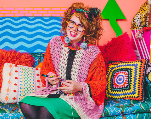 Individual wearing brightly colored crochet garments sitting in a brightly designed room.