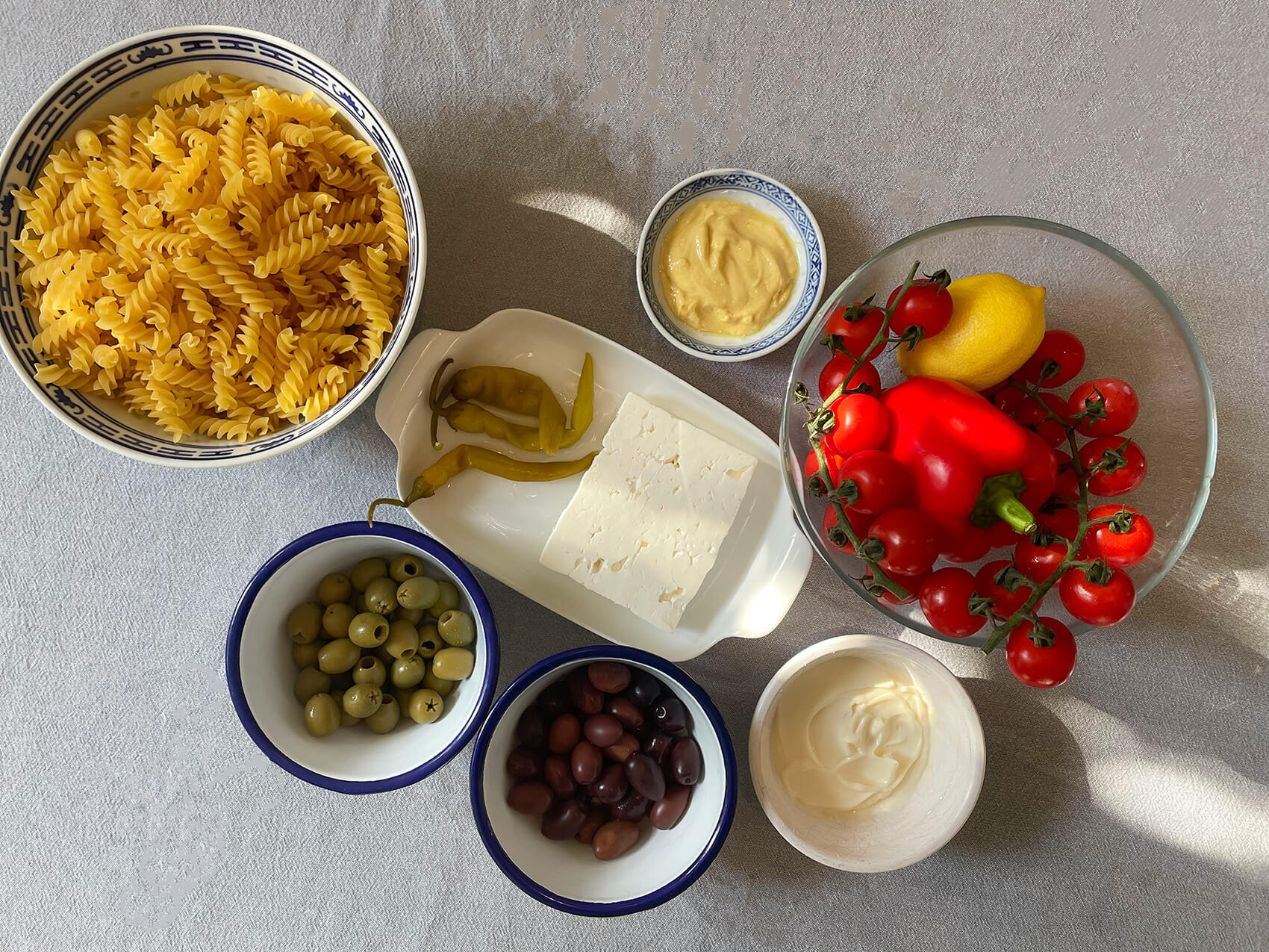 ingredients for a greek pasta salad, fusilli, feta cheese, kalamata olives, green olives, red peppers, mayo, mustard, tomatoes and lemon 