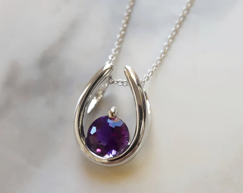 A 14k white gold pendant in an upright horseshoe shape, with a singe round Arizona amethyst at the bottom center of the curve. 