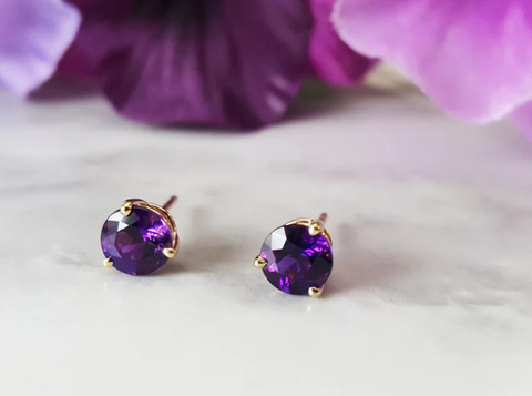 A Pair of small round purple amethyst stud earrings on a white background in from of purple flowers. 