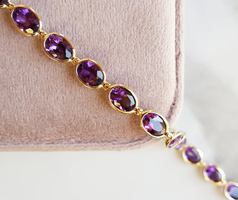 A bezel set Arizona Amethyst bracelet with oval shaped gemstones set in smooth yellow gold, The bracelet is laying across a soft light purple jewelry box 