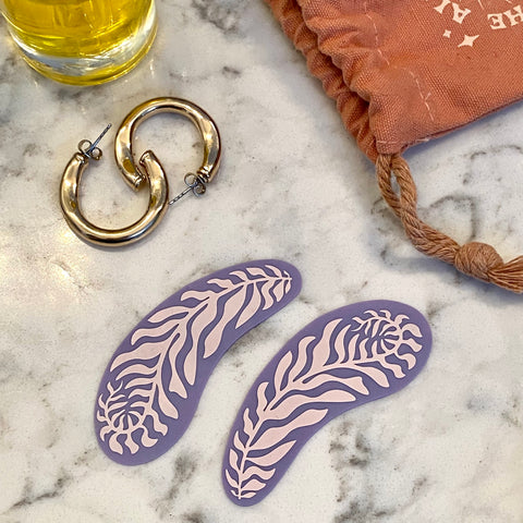 A picture of periwinkle eye masks with a fern pattern on a marble background next to gold hoop earrings