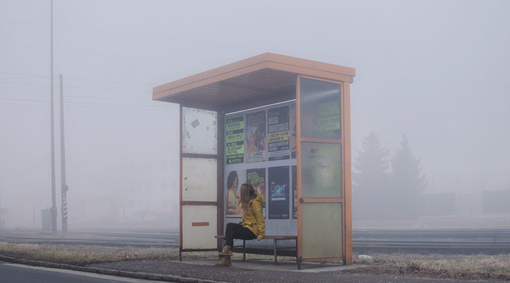 Eine Person mit gelber Jacke sitzt in einer Bushaltestelle, die von Nebel umhüllt ist.