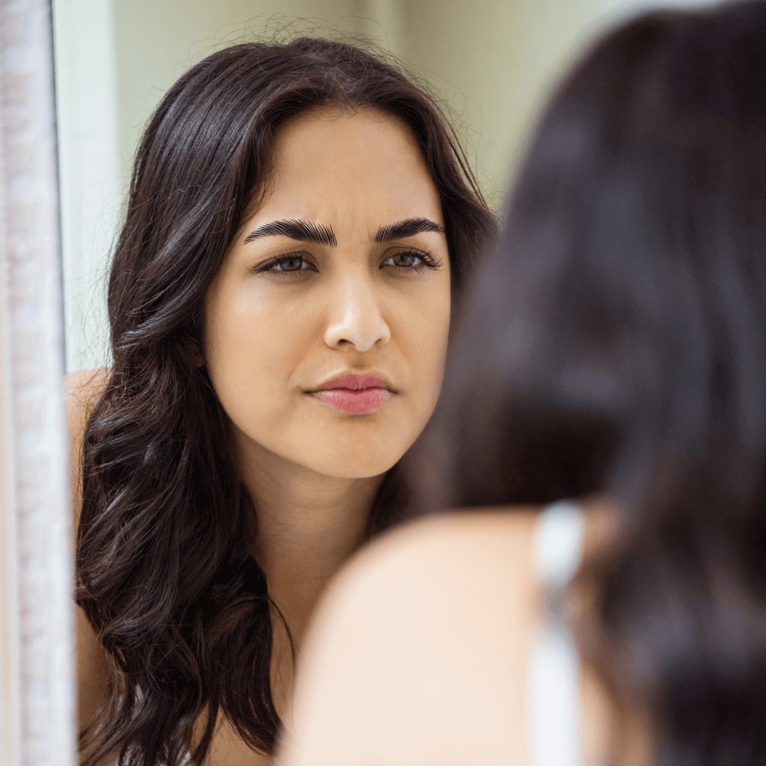 Woman with microblading looking into a small hand mirror with a concerned look on her face.