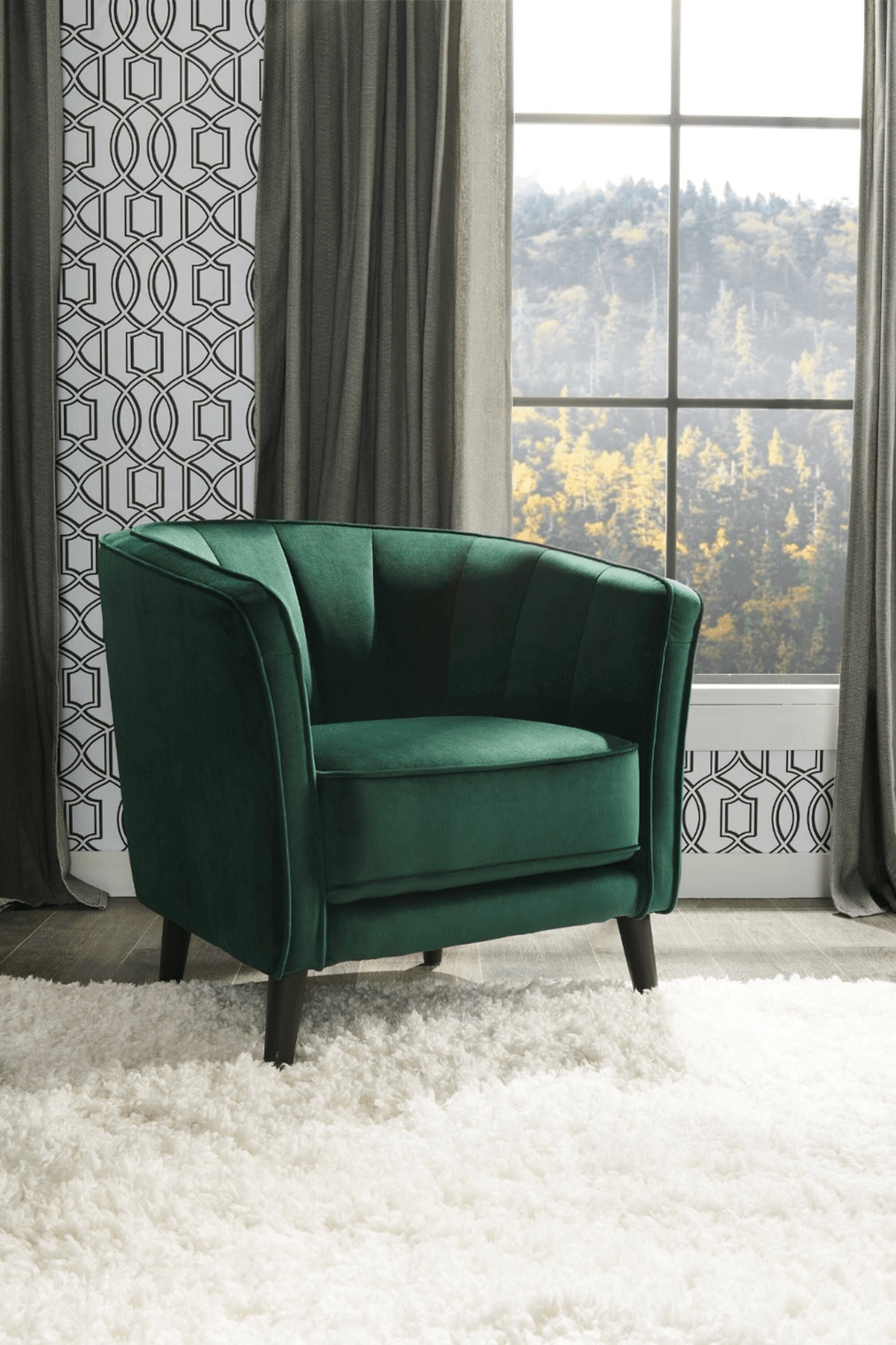 A photo of an accent chair in front of a window. The accent chair is upholstered with emerald green velvet fabric and looks retro with vertical line-tufting and dark wooden legs. The floor beneath the chair is covered with a white shag-carpet, and the wall in the background features a white patterned wallpaper and grey curtains. Through the window a forest is visible.