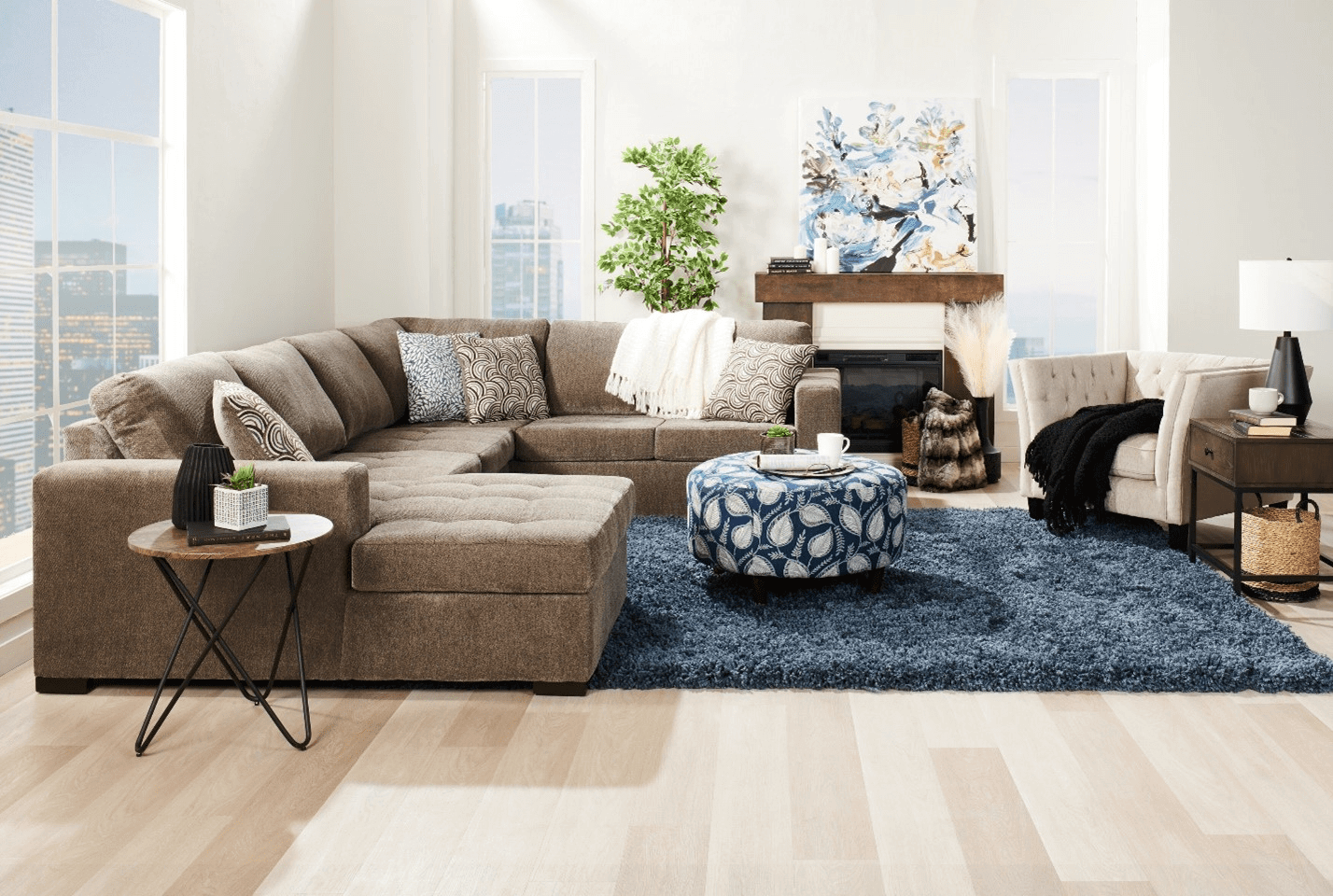 An image of a bright contemporary styled living room. A greyish brown chenille-upholstered sectional stands on a dark blue shag-carpet, paired with a blue accent ottoman. To the right stands a white accent chair with an industrial wooden side table. In front of the sectional is another side table, this one a light brown with modern black metal legs. In the background, a fireplace with a painting on the mantle is visible.