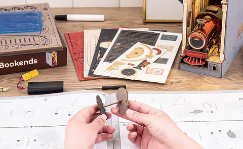 Two hands assembling wooden pieces of 'The Temporal Terminal' book nook kit.