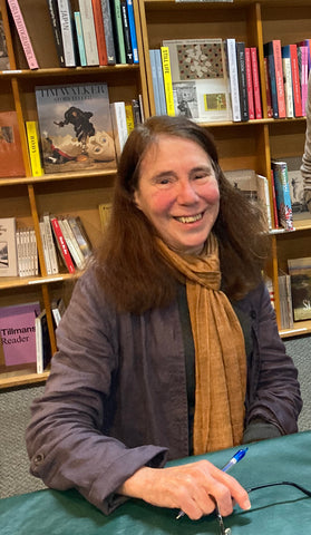 A woman with long dark hair sits at a table with bookshelves behind her. She is smiling.