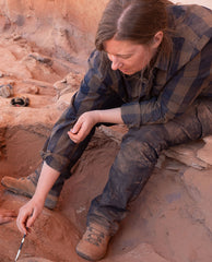 A young woman with light brown hair wearing brown-and-blue checkered shirt and jeans covered in dirt, is shown excavating with a trovel. She is shown seated on the edge of the trench and looking down.