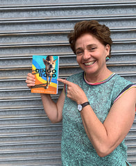 A photo of Rowena Lennox standing in front of a metal door and holding a copy of her book Dingo Bold. 