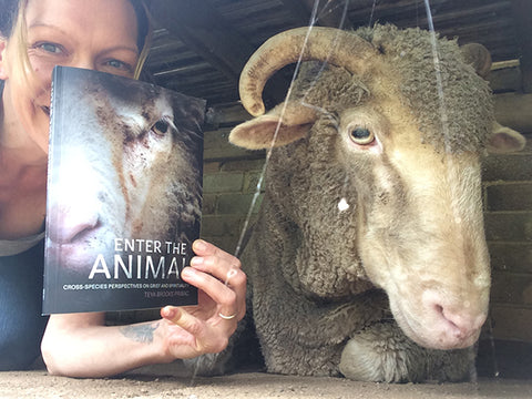 Photo of the heads of Teya and Orpheus-Pumpkin, the sheep. Teya is holding her book Enter the Animal with a photo of sheep's head on the cover. The book obscures most of Teya's head, leaving eyes and the arm visible.