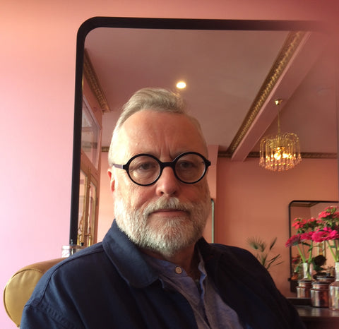 A close-up photograph of a man with white hair and a beard, wearing glasses with round black frames, sitting in a soft pink room.