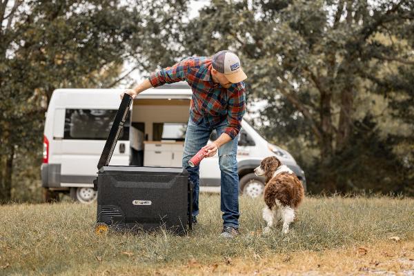 A man takes a cola and opens a BougeRV portable camping fridge with a dog beside him