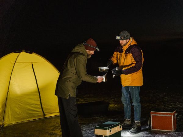 Two men dressing up in layers and sharing food and drinks while tent camping at night
