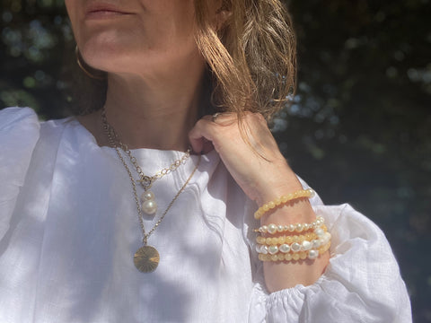 Image of jewellery modelled against a white linen dress, beaded and pearl bracelets as well as layered necklaces in gold vermeil with a round hammered as well as a freshwater pearl pendant 
