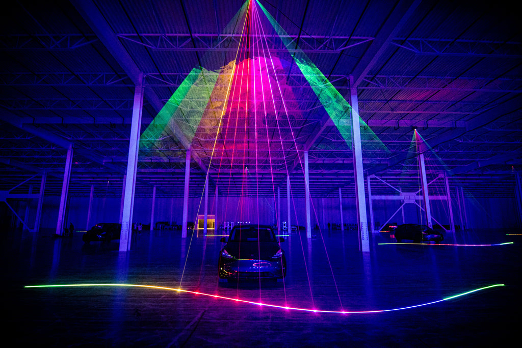 Multi-colored laser beam projects on to floor in front of Tesla vehicle.