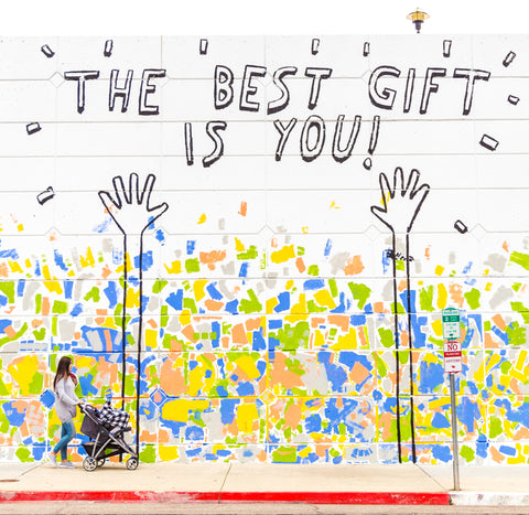 Photo of woman with a stroller walking by a colorful graphic mural with text that reads: "THE BEST GIFT IS YOU!"