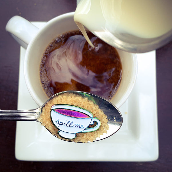 "Spill me" rainbow anodized teacup pin, in a spoonful of sugar with coffee & cream in the background.