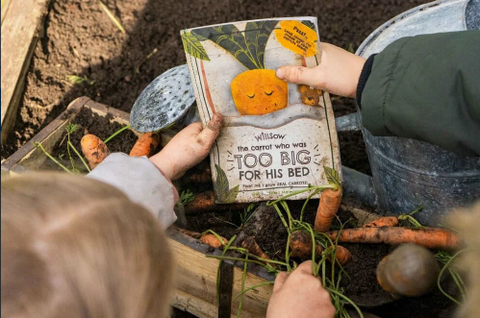 Children holding the "Plantable Carrot Children's Book" aver a carrot bed ready for planting