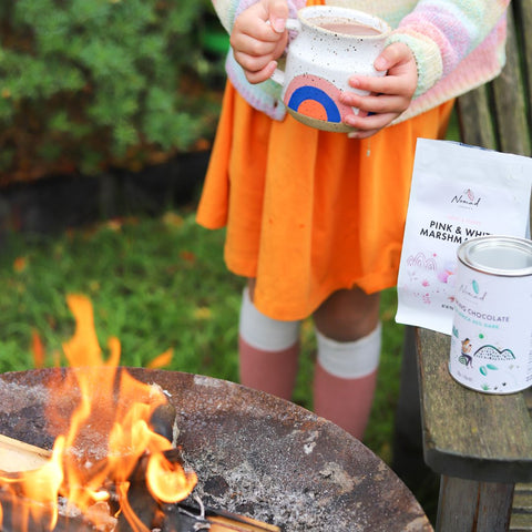 image of a girl holding cup with hot cacao and standing next to the fire pit with nomad hot chocolate west africa 45% dark and marshmallow and 