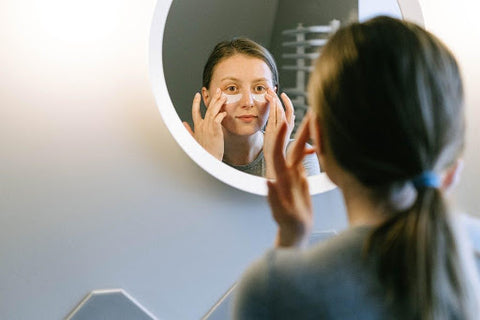 woman looking in mirror while putting skin cream under eyes