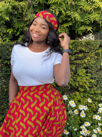 Ruth is a black woman with long straight black hair. She is wearing a white top with a red African wax print circle skirt and a matching headband.
