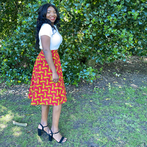 Ruth is a black woman with long straight black hair. She is wearing a white top with a red African wax print circle skirt and black strappy shoes. 