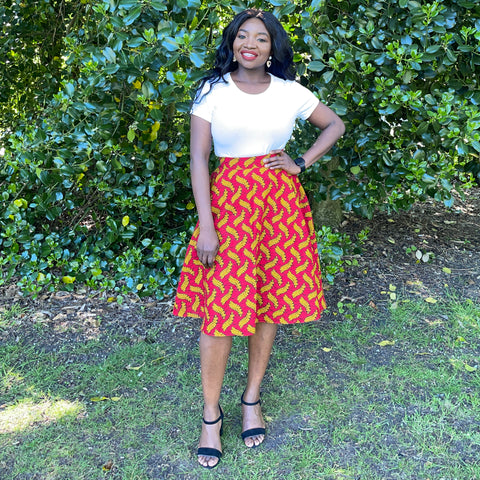 Ruth is a black woman with long straight black hair. She is wearing a white top with a red African wax print circle skirt and black strappy shoes. 