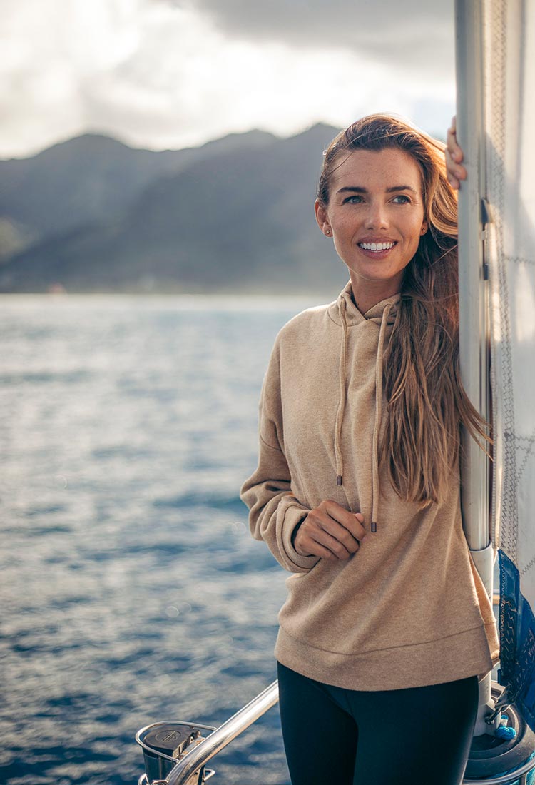 Rachel Moore sailing on her boat among blue skies, wearing a beige MPG hooded sweatshirt and blue leggings