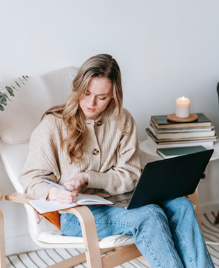 A woman writing in a notepad with a laptop on her knee and a candle lit in the background. Source: Pexels